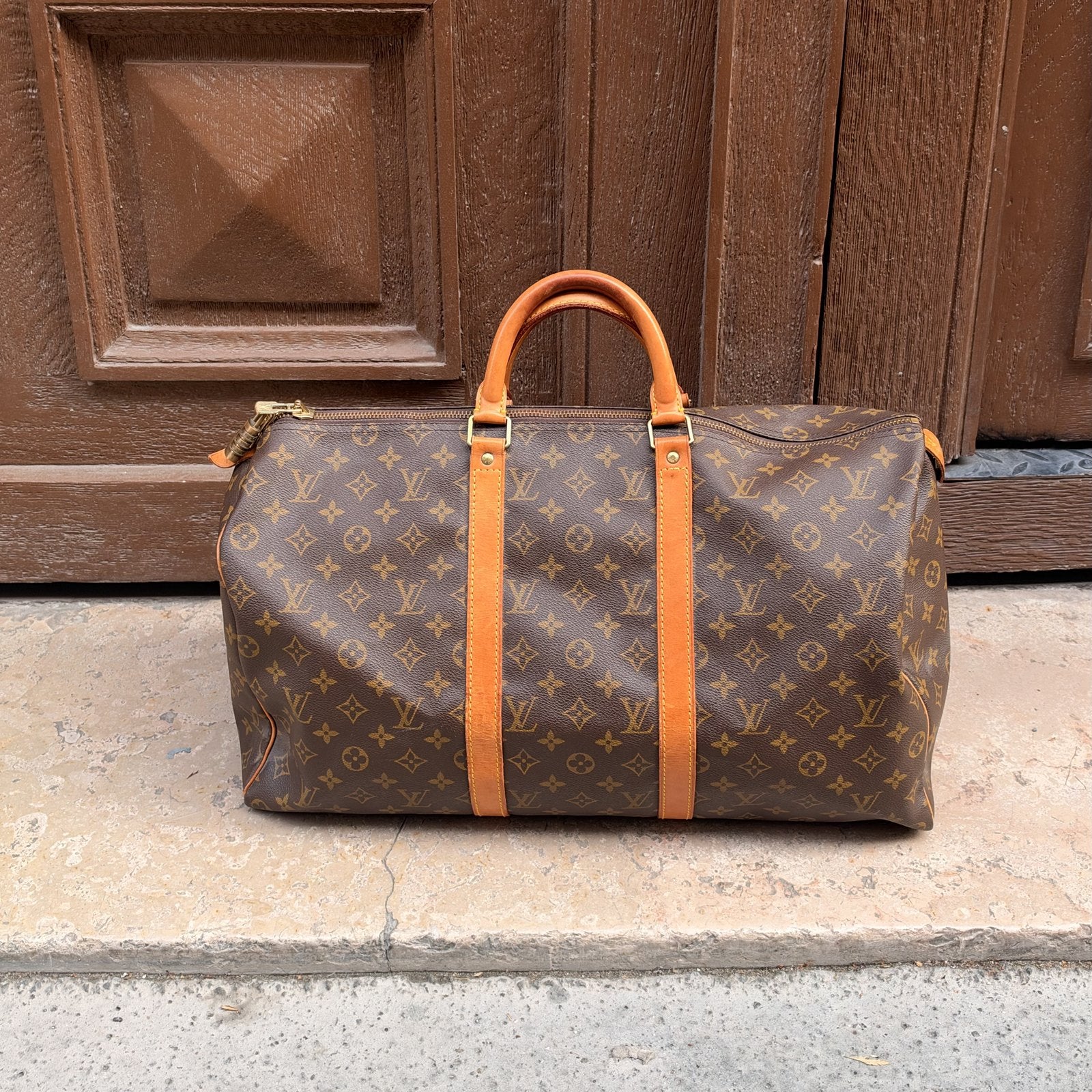 Sac de voyage Louis Vuitton Keepall en toile Monogram, anses patinées, posé sur une marche en pierre devant une porte en bois